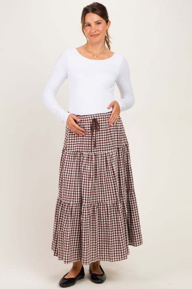 woman wearing a white long sleeve maternity top and a brown gingham tiered maternity skirt, holding her baby bump