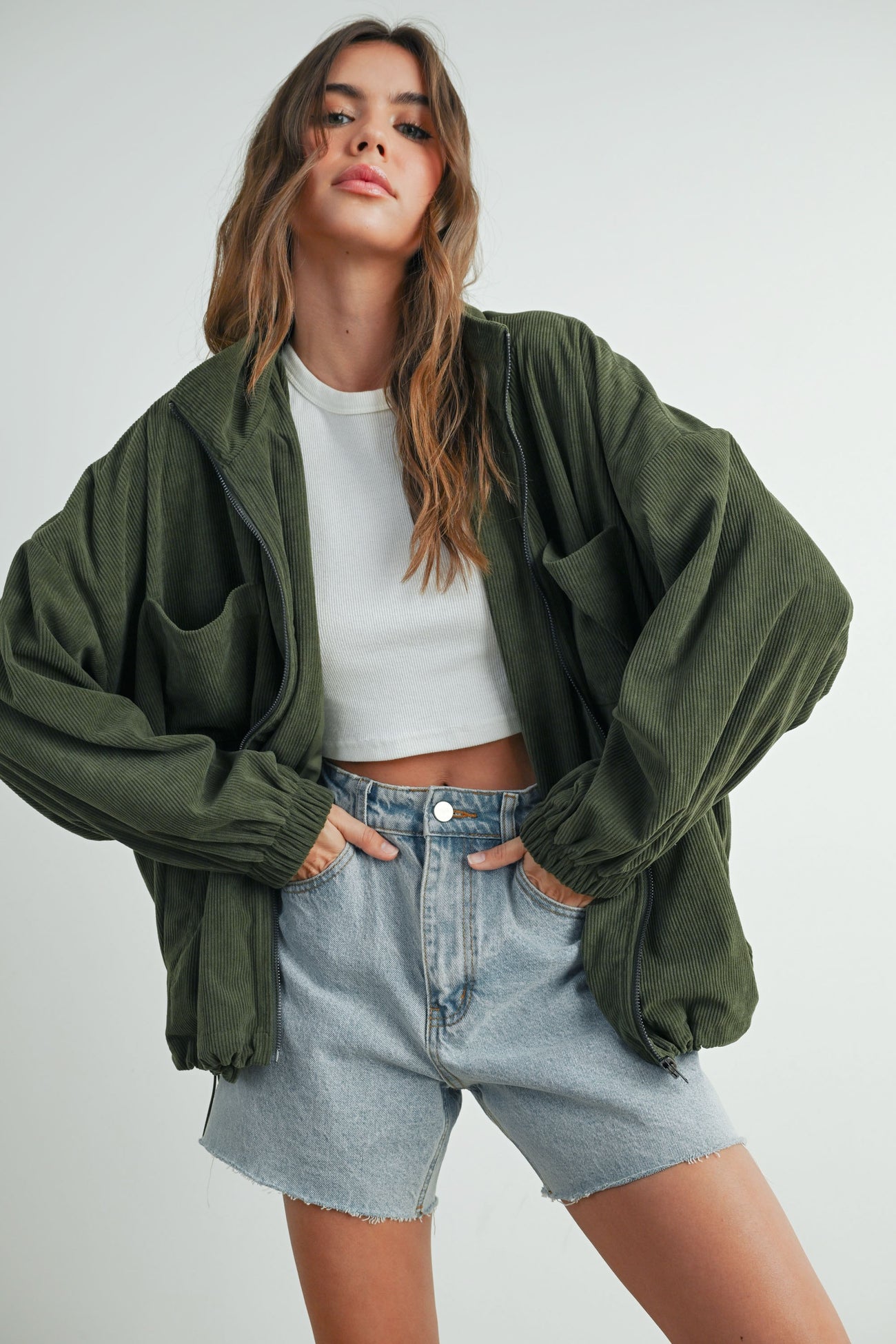 woman wearing an oversized olive green corduroy jacket over a white crop top and high-waisted denim shorts