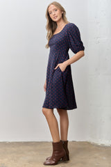 woman wearing a navy blue short sleeve dress with red polka dots and a square neckline, styled with brown ankle boots