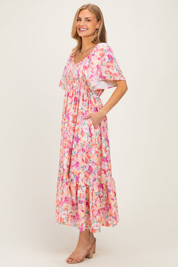 woman wearing a floral pink maxi dress with short sleeves and pockets, standing and smiling on a neutral background