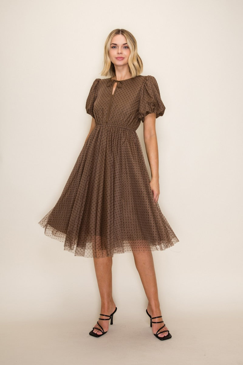 woman wearing a brown polka dot dress with puff sleeves and a keyhole neckline, standing against a neutral background