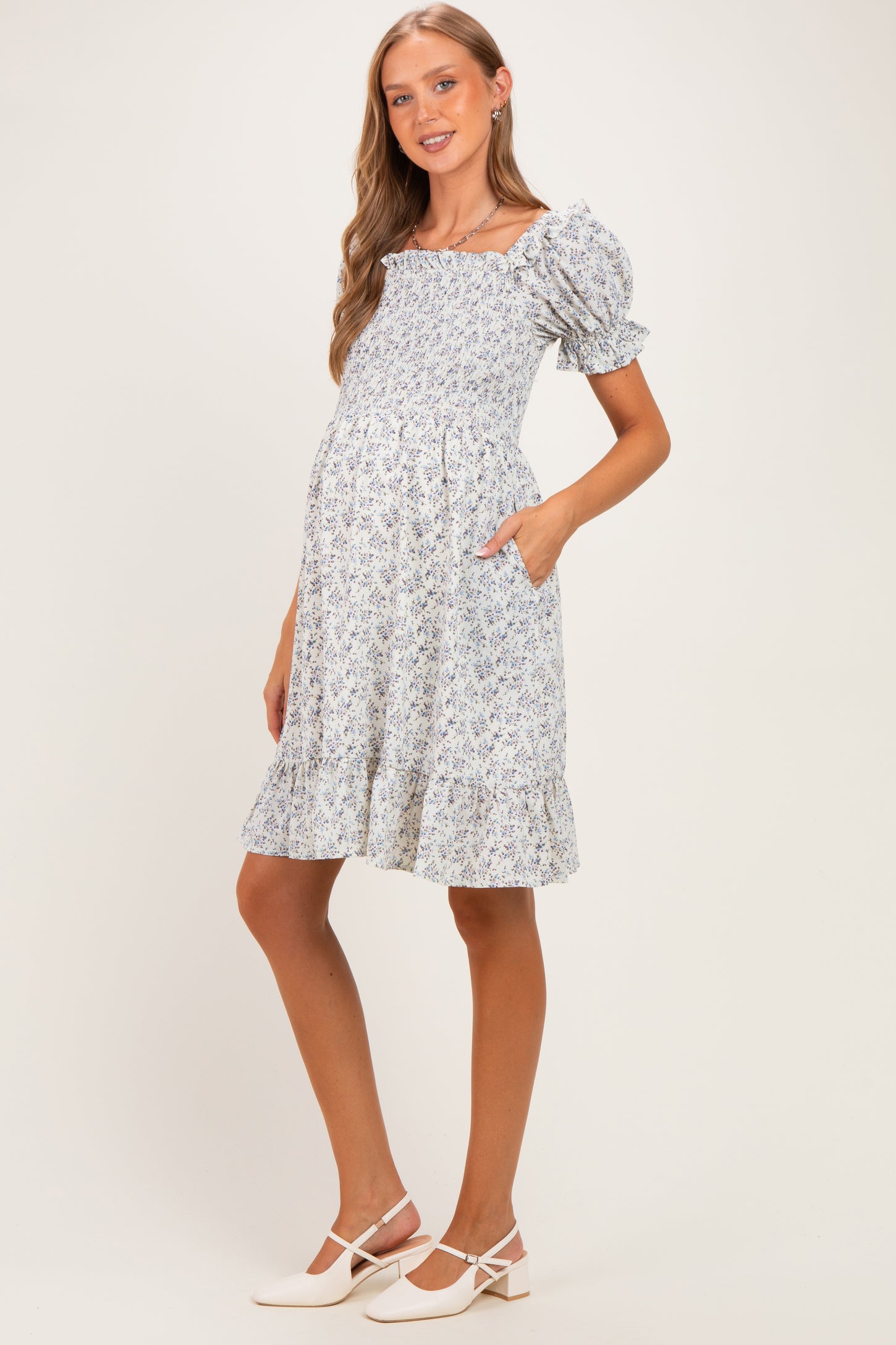 woman wearing a blue floral maternity dress with puff sleeves and ruffled hem, holding pockets, showing her baby bump