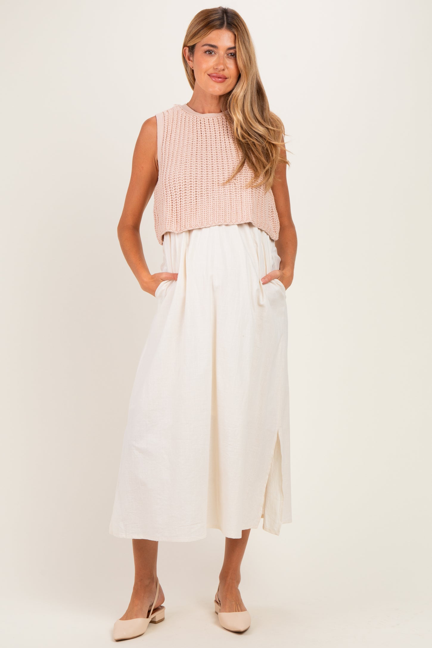 woman wearing a knitted blush maternity crop top over a white maxi dress with pockets and a side slit
