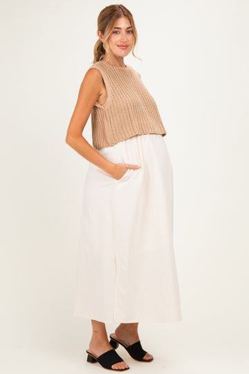 woman wearing a beige knitted crop top and a white long maternity dress with pockets, showing her baby bump