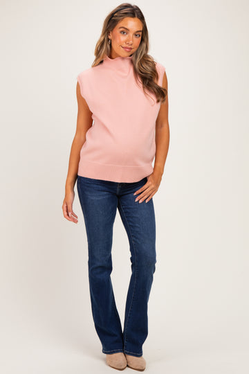 woman wearing a light pink sleeveless maternity sweater with a mock neck and blue flared jeans, showing her baby bump