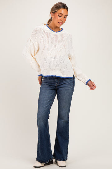 woman wearing a white laced maternity sweater with blue trim and blue flared jeans while standing and posing