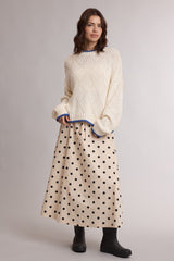 woman wearing a cream sweater with blue trim and a beige polka dot maxi skirt, paired with black ankle boots