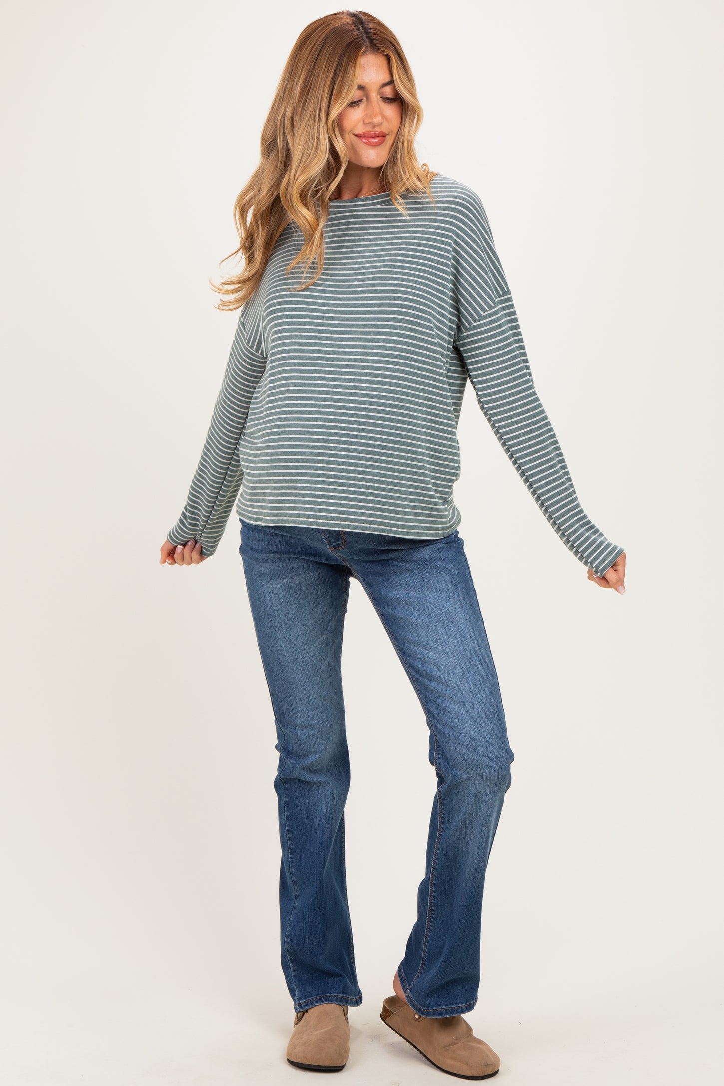 woman wearing a teal and white striped maternity top and blue flared jeans, smiling with a relaxed pose