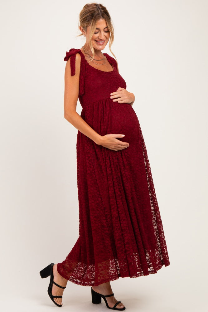 woman wearing a burgundy lace maternity maxi dress with tie shoulder straps, holding her baby bump