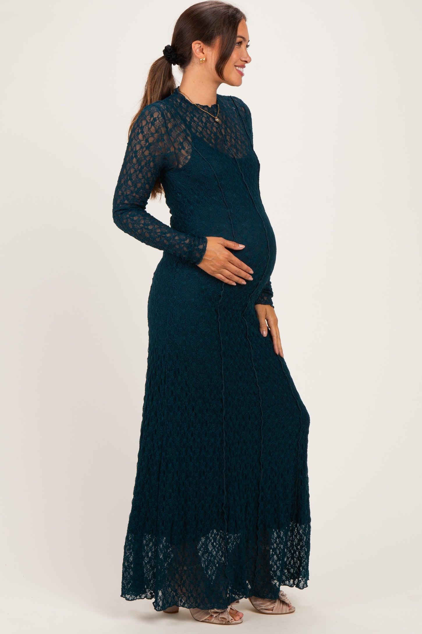woman wearing a teal lace long sleeve maternity gown with a flowing skirt and gently holding her baby bump