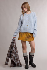woman wearing a light blue cable-knit cardigan over a mustard polka dot mini skirt and black rain boots, holding a plaid jacket