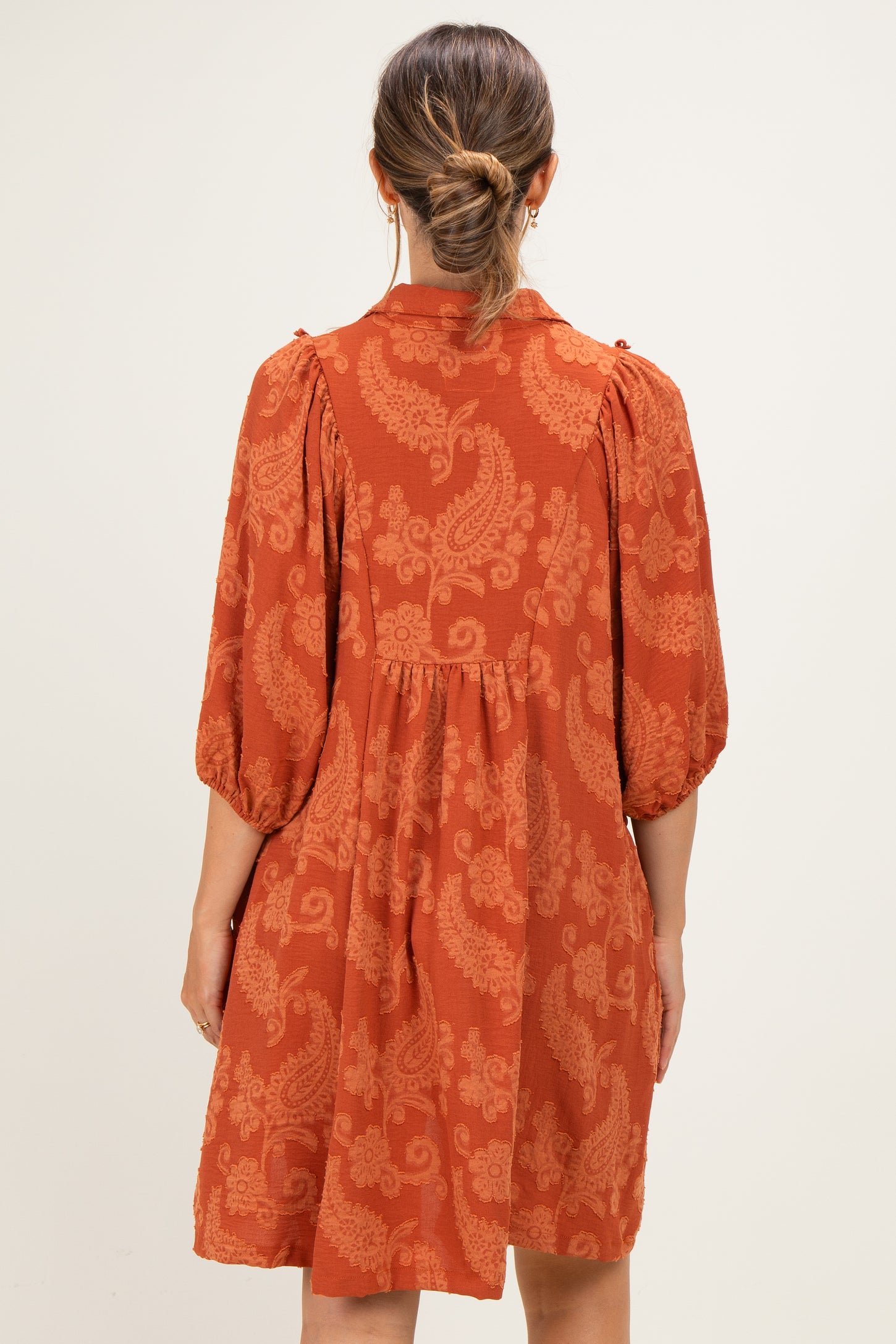 woman wearing a rust-colored maternity dress with puff sleeves and paisley print, showing back view