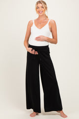 woman wearing a white lace trim tank top and black wide-leg maternity pants, gently cradling her baby bump