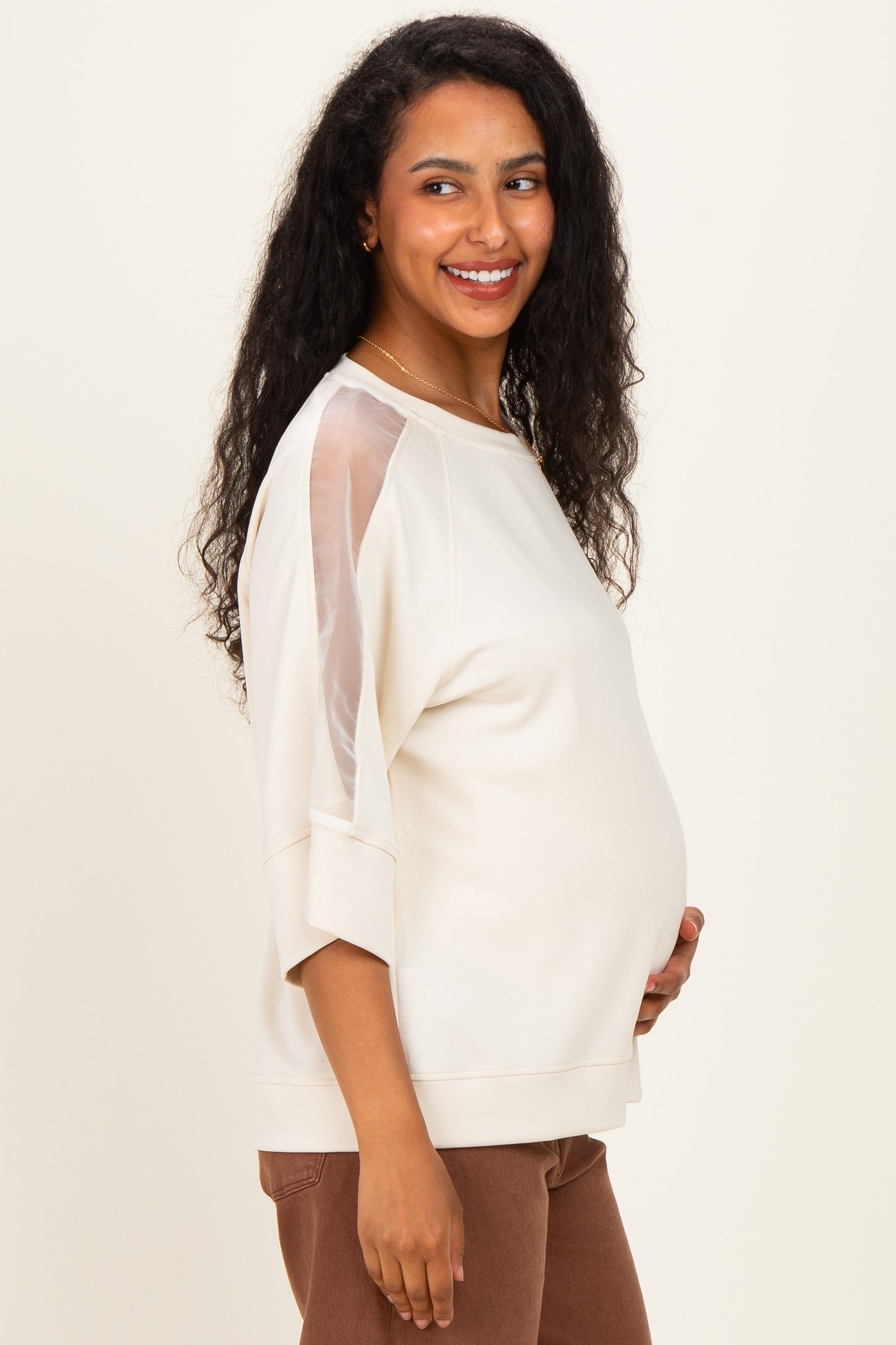 woman wearing a cream maternity top with sheer sleeves and a visible baby bump, smiling and standing sideways