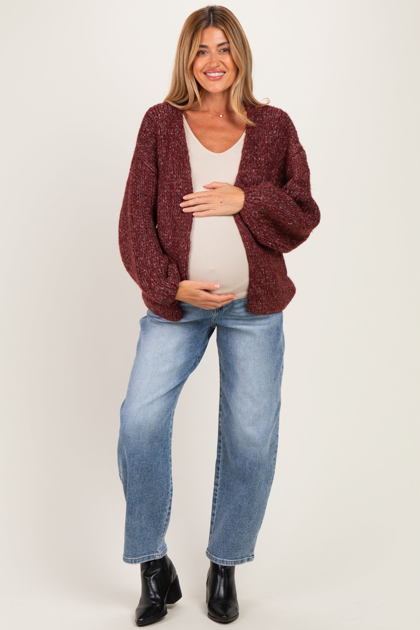 woman wearing a burgundy chunky knit maternity cardigan and light tank top, cradling her baby bump while standing in jeans