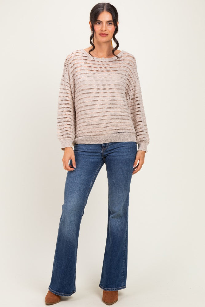 woman wearing a light brown open knit sweater and blue flared jeans, standing with a neutral background