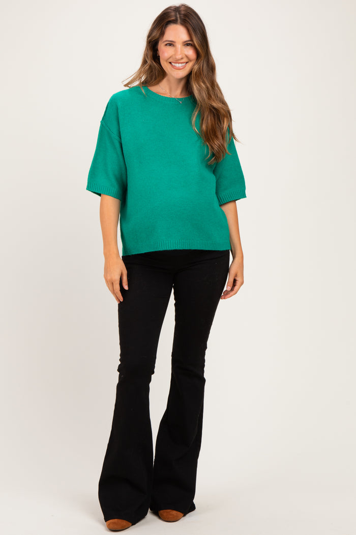 woman wearing a green oversized maternity sweater with short sleeves and black flared pants, showing her baby bump