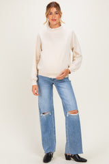 woman wearing a cream maternity sweatshirt and distressed light blue jeans, gently cradling her baby bump