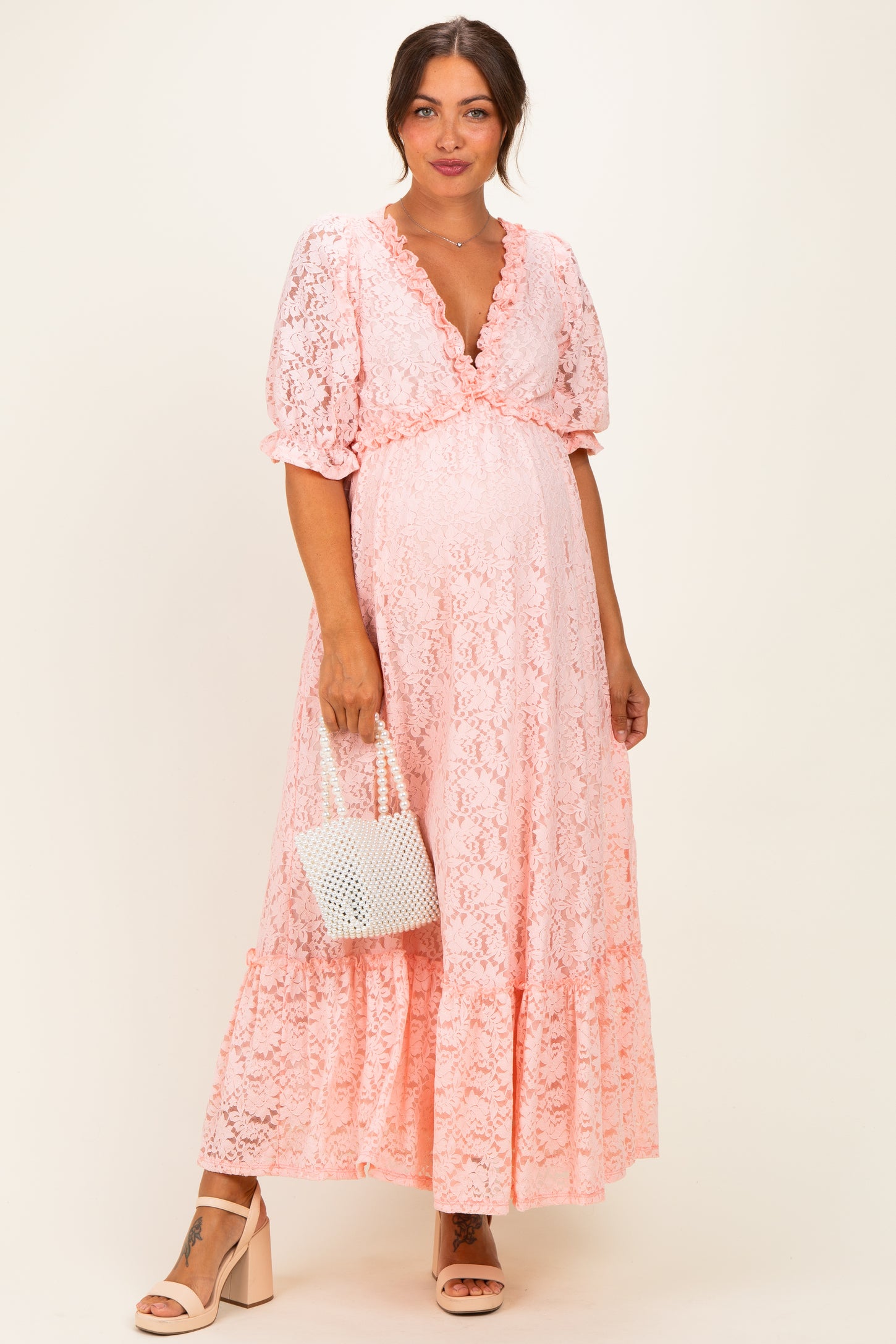 woman wearing a pink lace maternity dress with puff sleeves and ruffled hem, holding a white handbag and showing her baby bump
