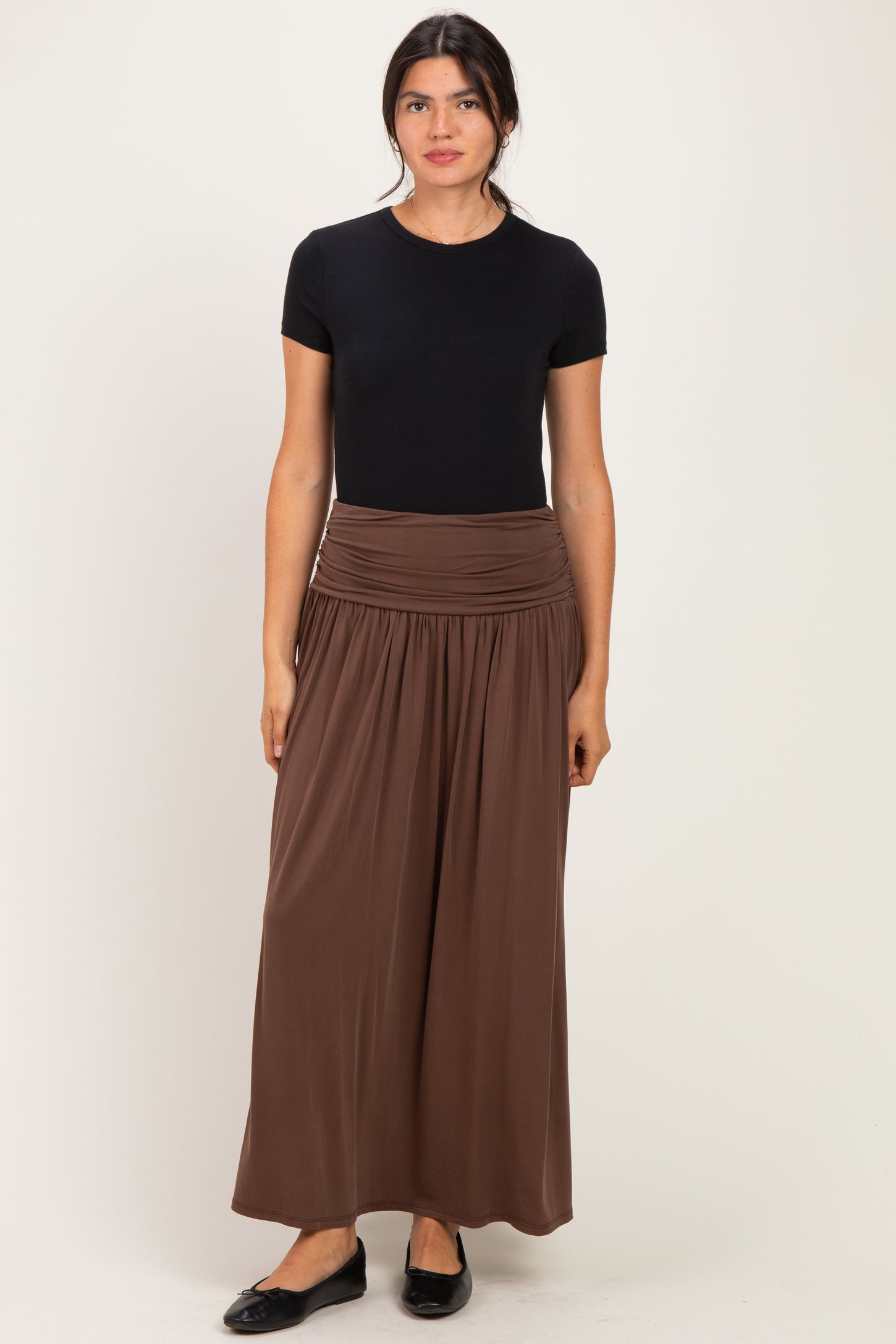 woman wearing a black short sleeve top and a brown maxi skirt, standing against a neutral background