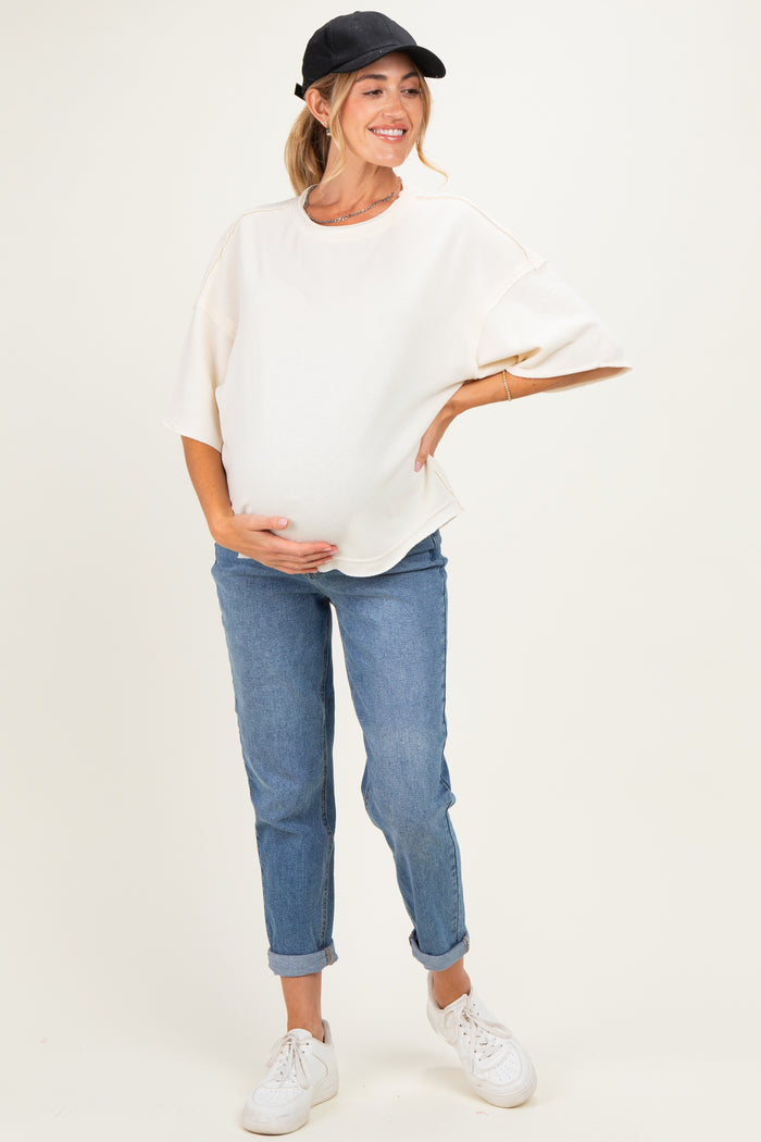 woman wearing a cream oversized maternity top and blue jeans, holding her baby bump while smiling.
