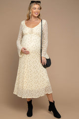 woman wearing a cream floral lace maternity dress with a baby bump and bell sleeves, holding her belly with a black purse and boots