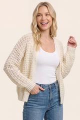 woman wearing a cream open-knit cardigan over a white tank top and high-waisted jeans, smiling at the camera