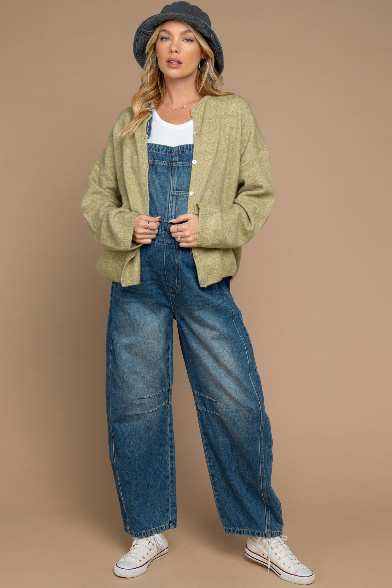 woman wearing a light green cardigan over a white tank top and blue denim overalls, styled with a gray hat