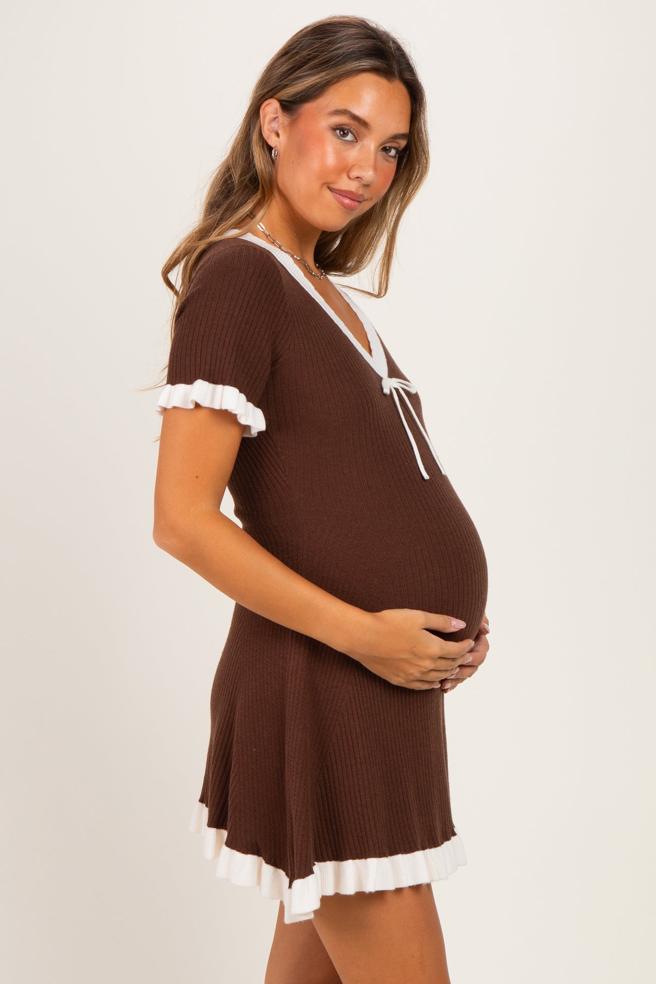 woman wearing a brown ribbed maternity dress with white trim and a tie detail, holding her baby bump