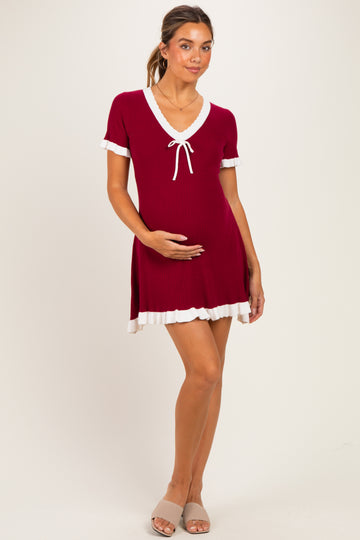 woman wearing a burgundy short sleeve maternity dress with a tie detail and white ruffled hem, holding her baby bump