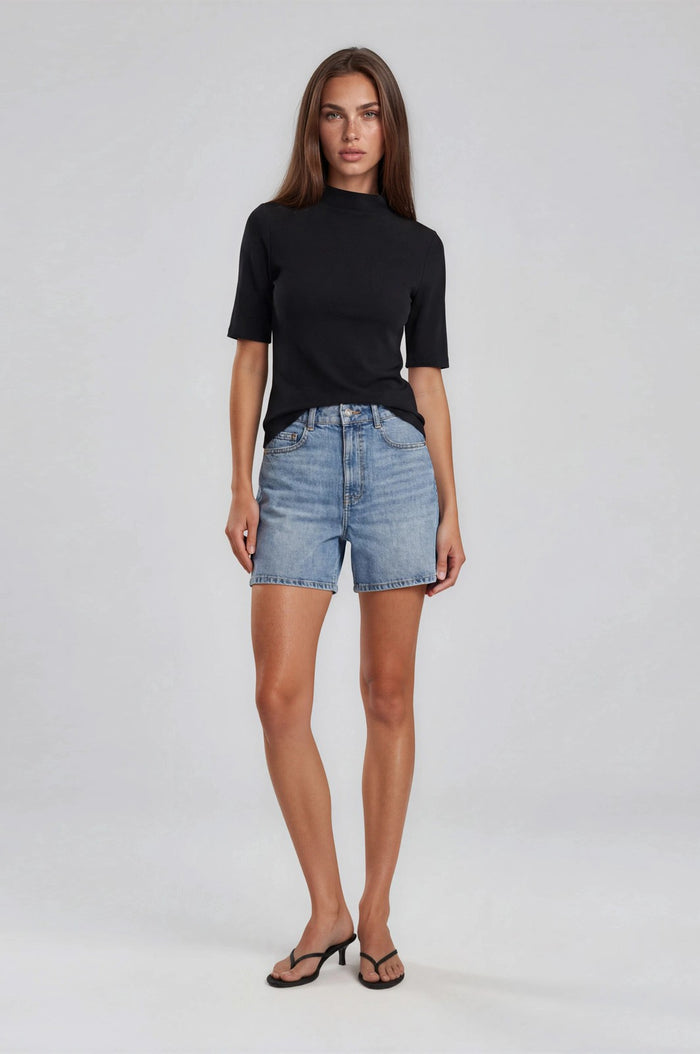 woman wearing a black short sleeve top and light blue denim shorts, standing against a neutral background