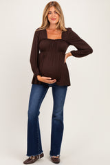 woman wearing a brown long sleeve maternity top and blue flared jeans, holding her baby bump