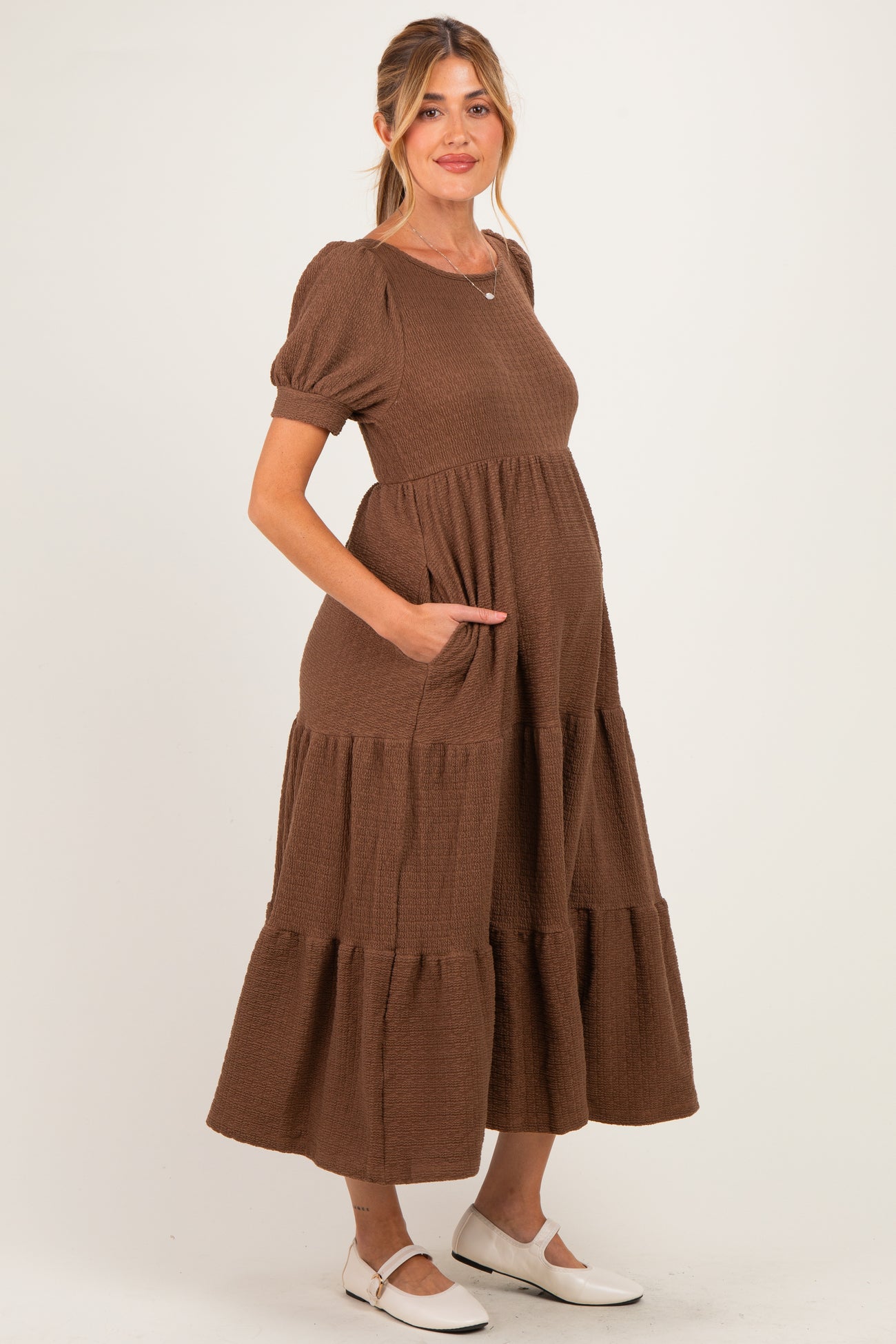 woman wearing a brown tiered maternity dress with short puff sleeves and pockets, showing her baby bump