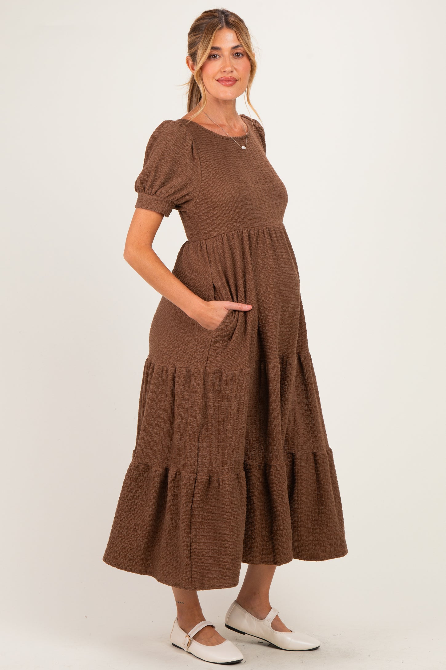 woman wearing a brown tiered maternity dress with short puff sleeves and pockets, showing her baby bump