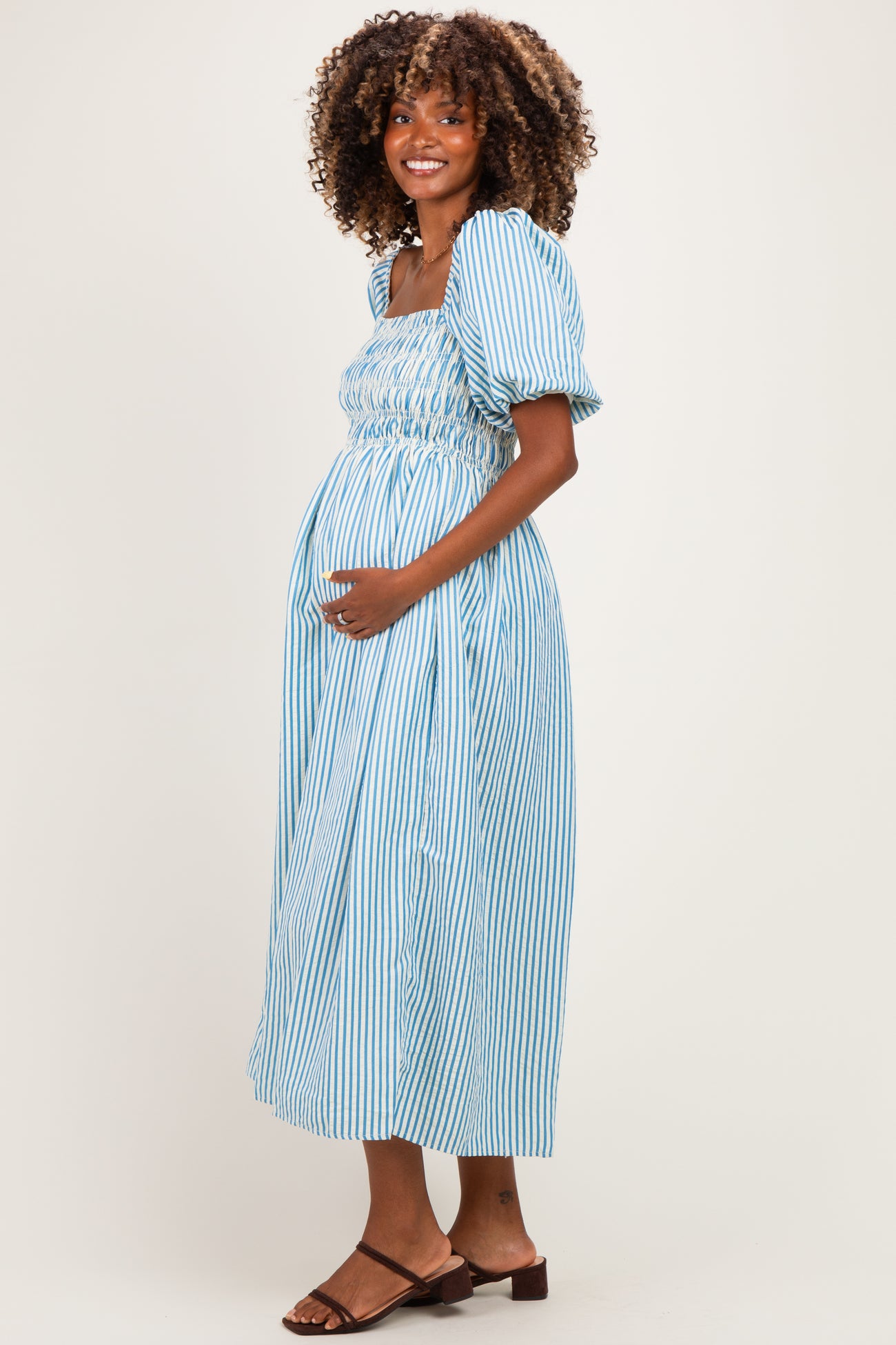 woman wearing a light blue striped maternity dress with puff sleeves and smocked bodice, holding her baby bump