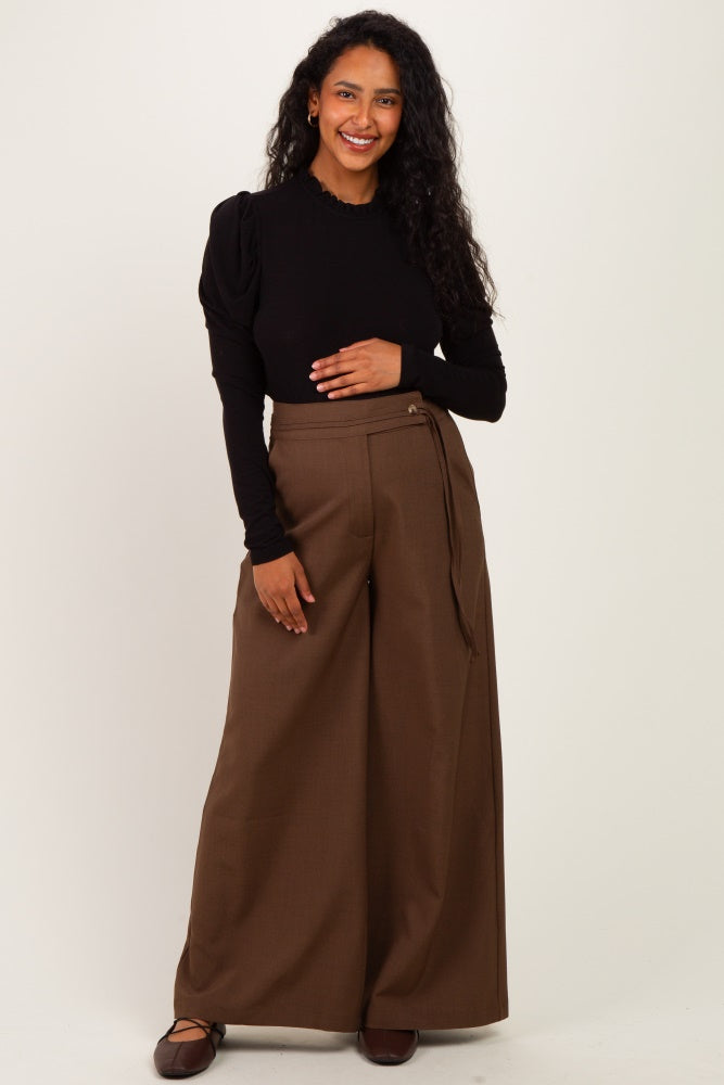 woman wearing a black long sleeve top and brown wide leg maternity pants, holding her baby bump