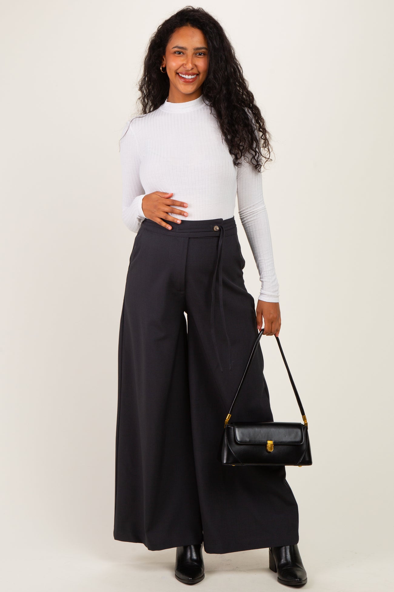 woman wearing a white long sleeve fitted top and high-waisted black wide-leg maternity pants, holding a black handbag
