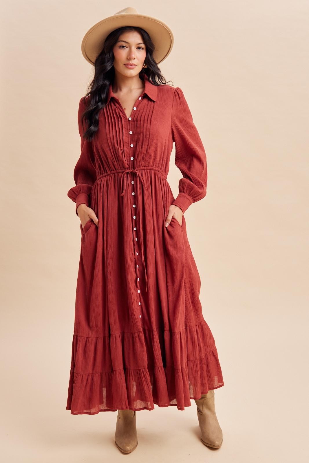 woman wearing a rust long sleeve maxi dress with a button-up front, pockets, and ruffled hem, paired with a beige hat and boots