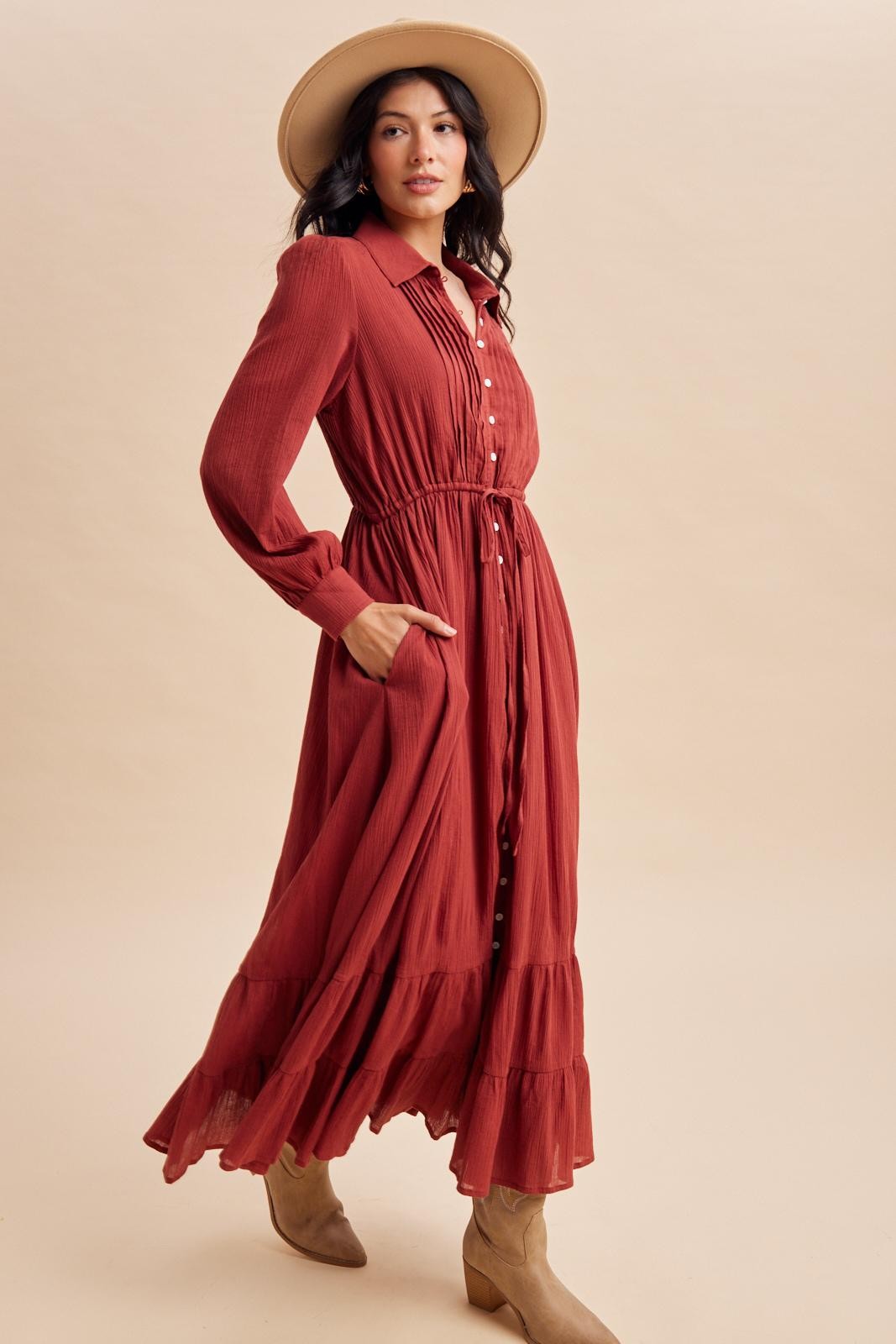 woman wearing a rust colored long sleeve button-up maxi dress with an elastic waist and ruffle hem, paired with a wide-brim hat and boots
