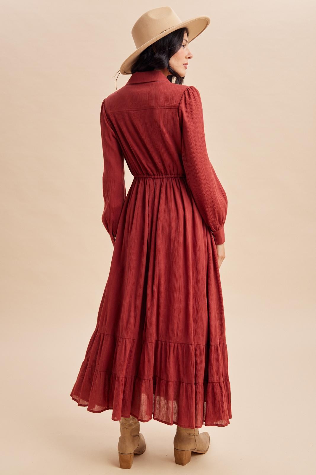 woman wearing a rust-colored maxi dress with long sleeves and a tiered skirt, standing with her back to the camera and wearing a hat