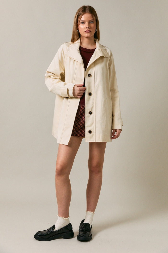 woman wearing a cream-colored button-up coat over a maroon top and plaid skirt, paired with black shoes and white socks