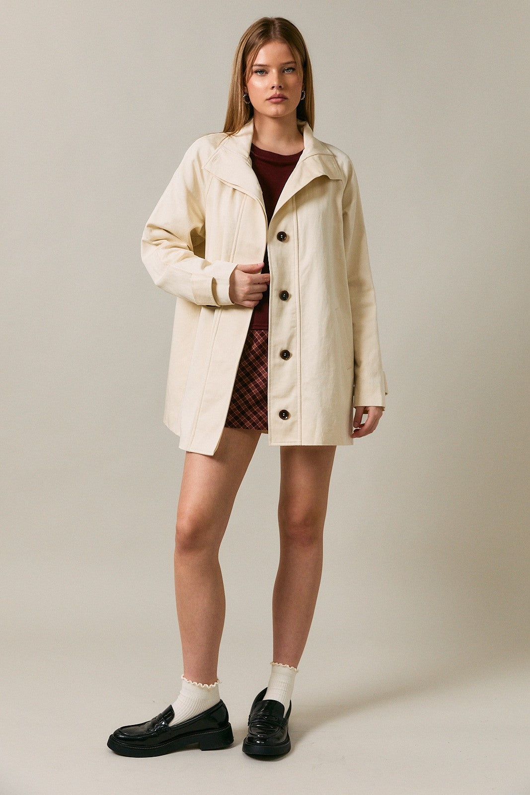 woman wearing a cream-colored button-up coat over a maroon top and plaid skirt, paired with black shoes and white socks