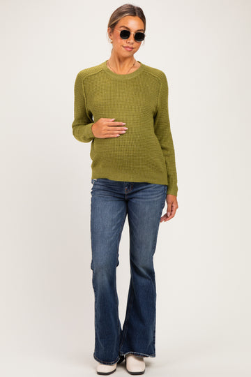woman wearing an olive green knit maternity sweater and flared jeans, holding her baby bump