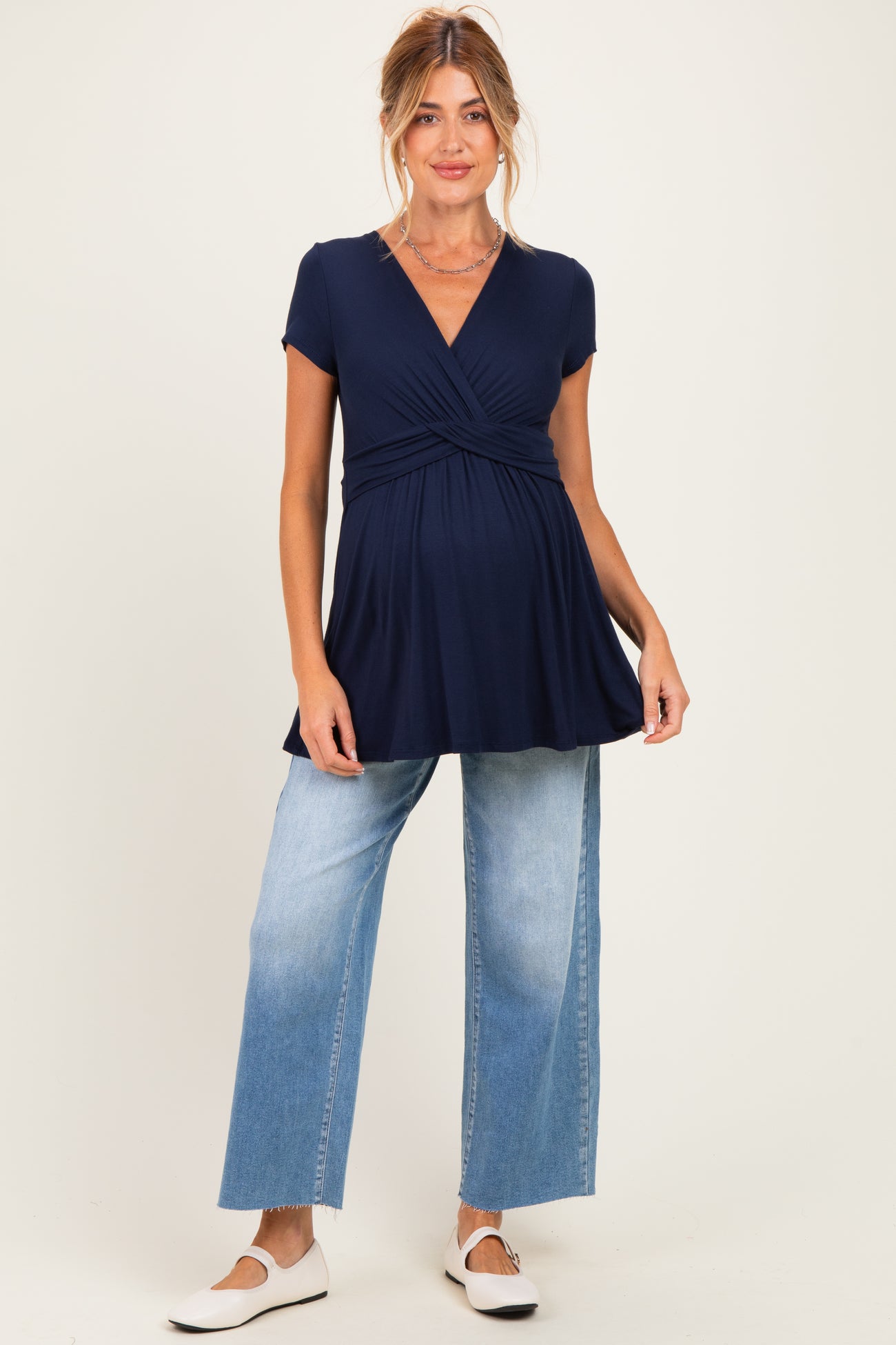 woman wearing a navy blue short sleeve maternity top with a twist design and light blue wide leg jeans, showing her baby bump
