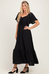 woman wearing a black maxi dress with flutter sleeves and a ruffled hem, styled with black ankle strap heels