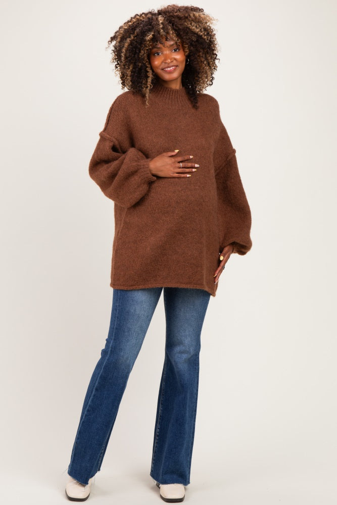 woman wearing a brown oversized maternity sweater and blue flared jeans, holding her baby bump