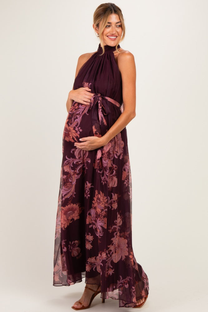 woman wearing a burgundy floral maternity maxi dress with a halter neck and a tie waist, cradling her baby bump