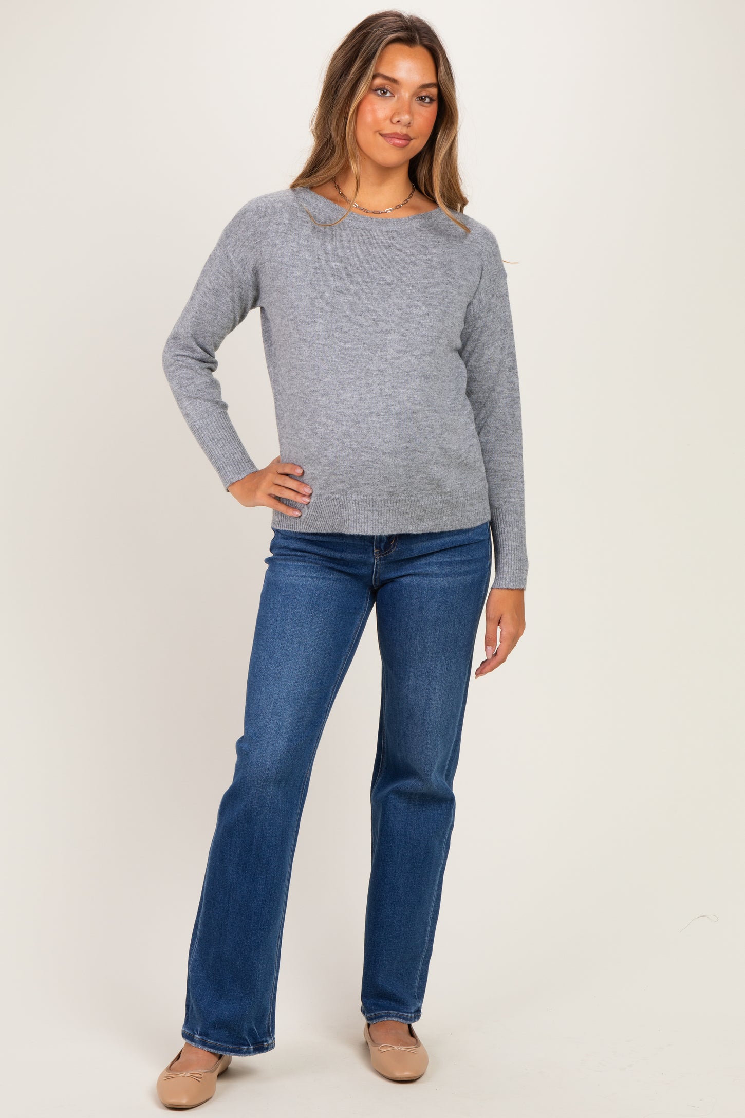 woman wearing a light gray long sleeve maternity sweater and blue flared jeans, with a subtle baby bump visible