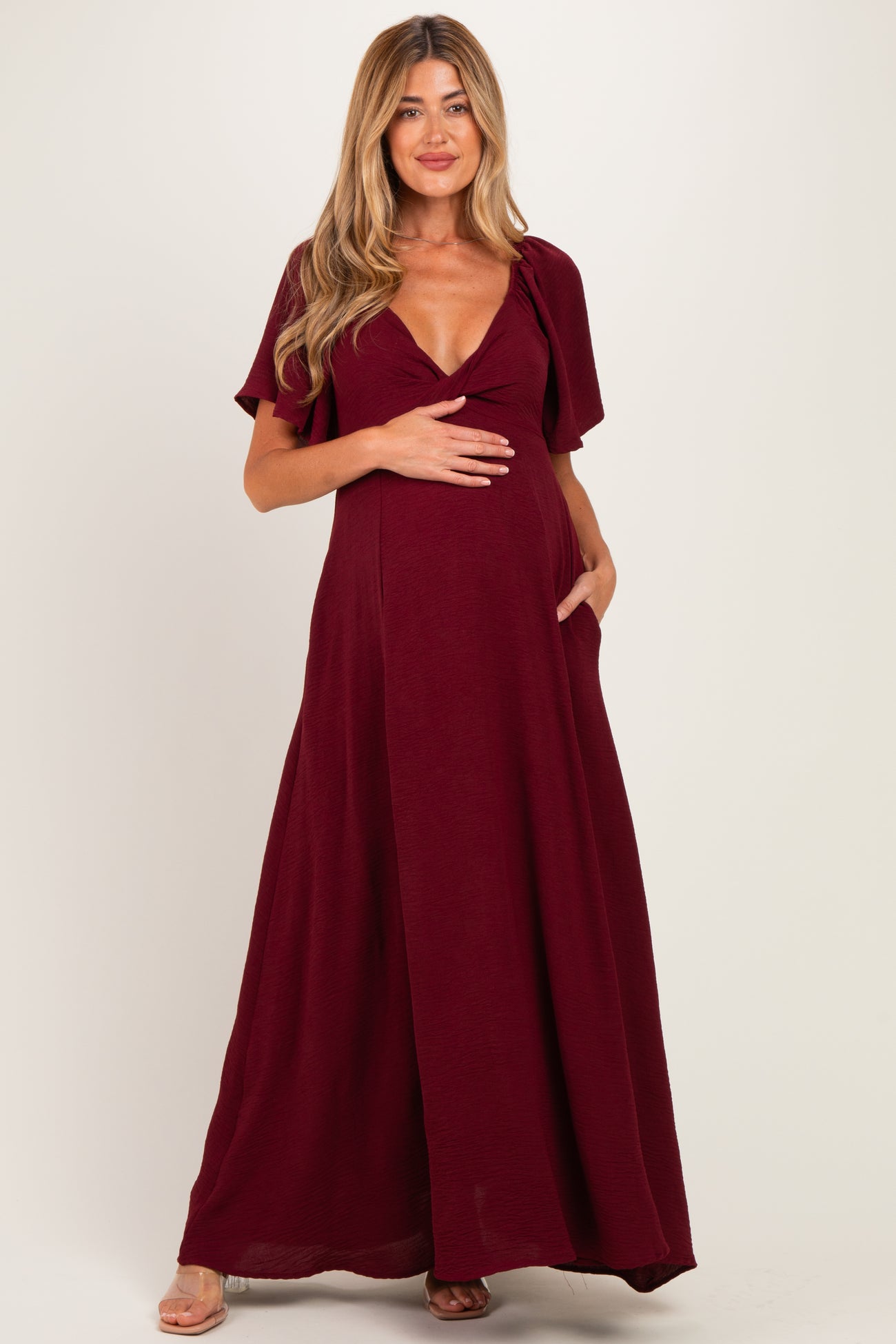 woman wearing a burgundy maternity maxi dress with short sleeves and a tie detail, holding her baby bump