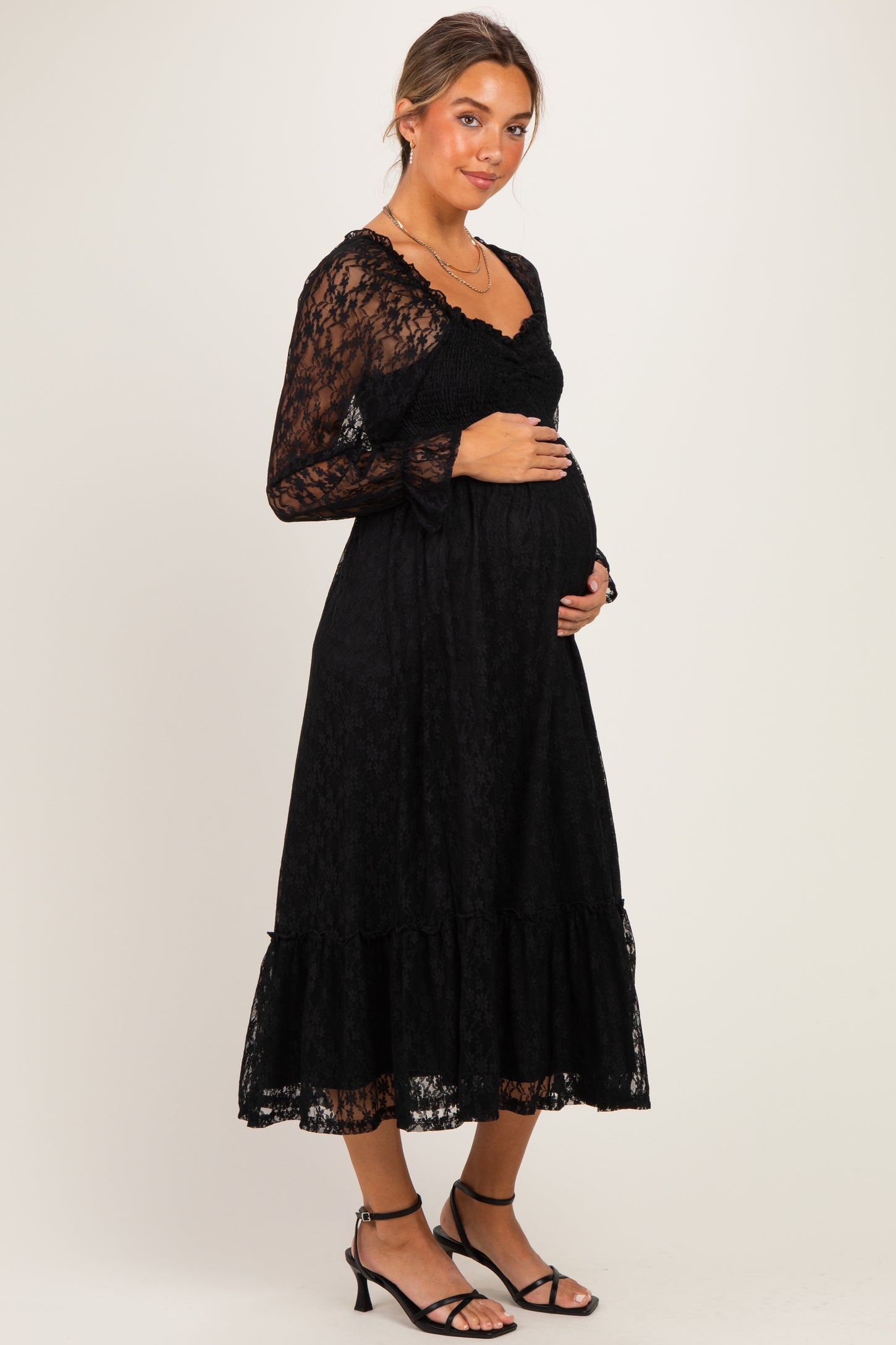 woman wearing a black lace maternity dress with long sleeves and a tiered skirt, cradling her baby bump